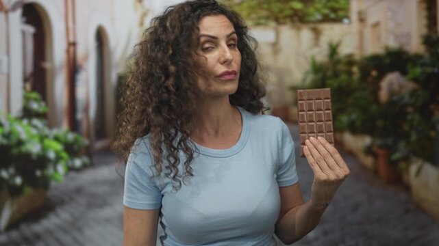 Young hispanic woman holds chocolate bar and points finger to bar on a sunny cobblestone city street; caution warning.