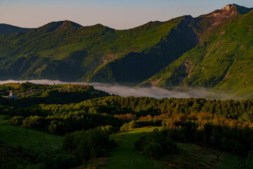 Naklejka premium Low fog ribbon beneath sunlit Caucasus slopes and forested hills