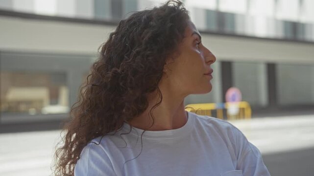 Woman with curly hair looks sideways showing face and closed lips under sunlight on street beside modern building facade; serenity.