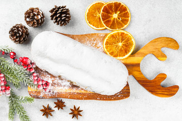 Classic Christmas Stollen covered with powdered sugar icing on wooden board with dried oranges, pine branches and cones on white textured background. Traditional holiday food, sweet fruit loaf