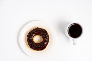 Top-down view of a chocolate-glazed doughnut with spherical sprinkles on a white plate, positioned beside a white cup of black coffee on a plain background.