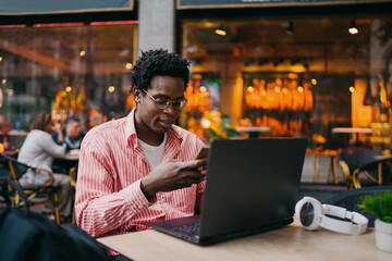 Young African businessman multitasking on phone and laptop in outdoor café, managing tasks on the go, representing mobile productivity and tech-enabled flexibility.