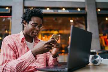 Smiling African freelancer pausing to look at camera while holding smartphone near laptop, expressing positive emotion and ease in tech-powered remote career.