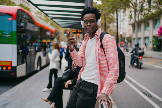 Black man smiling while raising smartphone camera for video or photo at bus stop, wearing headphones and backpack, illustrating mobile self-expression and tech comfort in freelance day