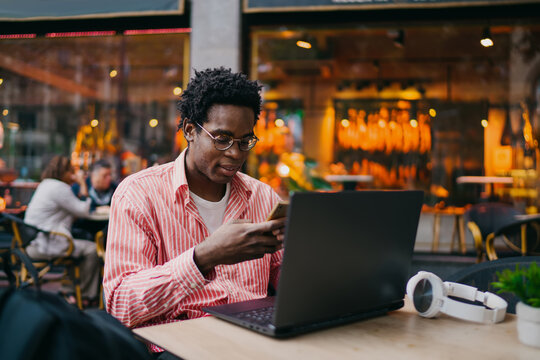 Young African businessman multitasking on phone and laptop in outdoor café, managing tasks on the go, representing mobile productivity and tech-enabled flexibility.