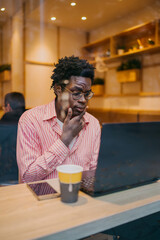 African man in café resting head on hand, eyes focused on screen, deep in thought while working on laptop, capturing modern tech fatigue, problem solving and solo freelance work.