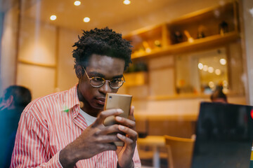 Focused African man in striped shirt holds smartphone with both hands, fully immersed in screen, portraying tech engagement and solo concentration in digital freelance routine.