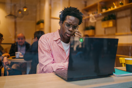 Young man on phone looks confused while using laptop, suggesting misunderstanding or tech issue. Reflects real-time troubleshooting in a freelance work scenario.