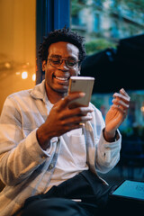 Confident young man seated at a cafe entrance, passionately speaking on a smartphone, expressing ideas while balancing work and conversation with his tablet on his lap.