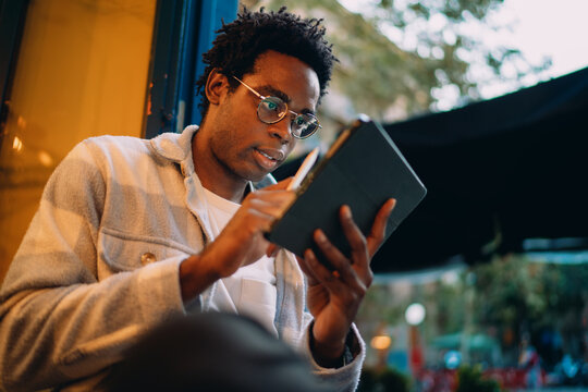 Side view of a man drawing or writing on a tablet with a stylus, sitting near a café window. Captures digital creativity and mobile design work in an open, flexible environment.