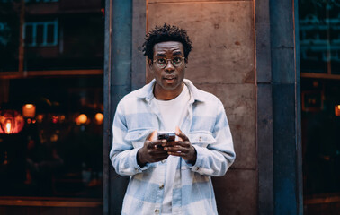 Surprised young man looking at camera, holding a smartphone, gesturing with one hand. Captures digital interaction, real‑time reaction and mobile‑first lifestyle in an expressive urban tech context.