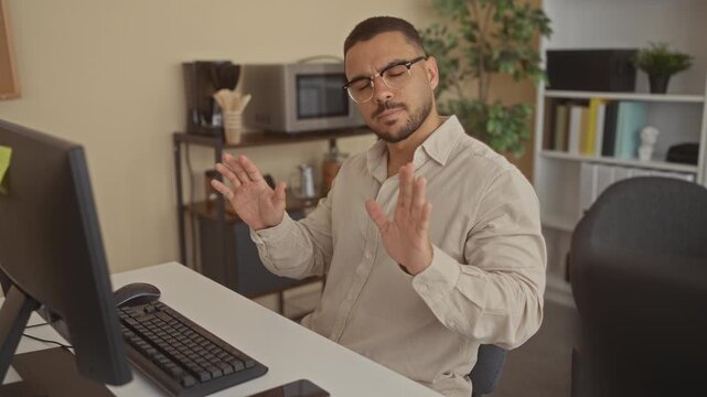 Man raising hands in a stop gesture at a white computer desk with keyboard and monitor in a bright office interior; refusal.