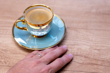 Hand resting near a luxurious blue-and-gold porcelain coffee cup on a wooden table. Classic decorative design suggesting comfort, relaxation and an elegant coffee moment.