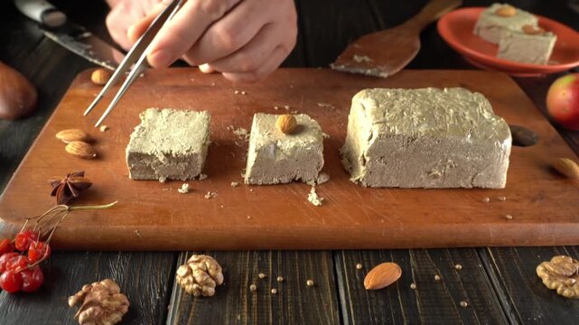A person cuts homemade halva on a wooden board, showcasing its texture. Surrounding the halva are nuts and fruits, creating a warm and inviting kitchen scene