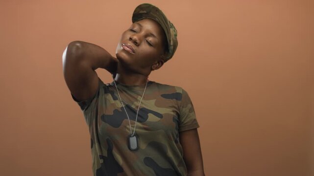 Young woman soldier wearing camouflage uniform, cap, and dog tags touches neck in peach studio; pride duty.