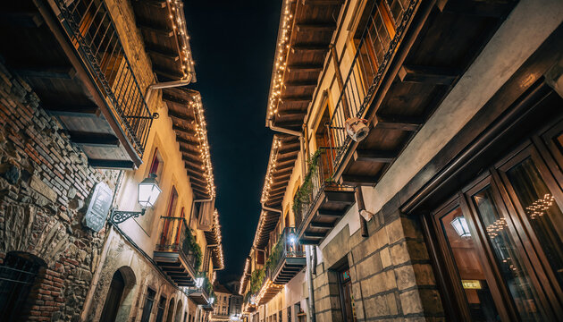 calle estrecha de un casco antiguo, iluminada con luces navide&ntilde;as c&aacute;lidas que bordean los tejados y balcones de edificios de piedra y madera, creando un ambiente festivo y acogedor durante la noche.