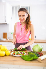 Positive woman is cutting vegetables for salad in kitchen at home