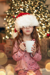 A little girl in a Santa hat holds a mug of hot drink next to the Christmas tree. Merry Christmas!