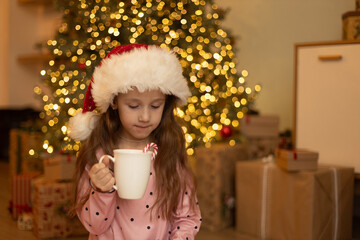 Little girl in pajamas holding mug with hot drink in decorated living room, winter weekend and Christmas holidays.