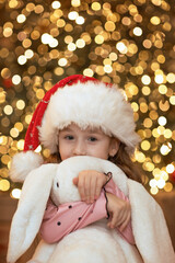 A cute child with a Christmas tree. A happy child in a red hat sits near the tree and hugs a toy.