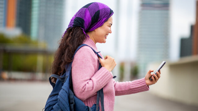 Young Asian woman using smartphone while smiling in urban setting