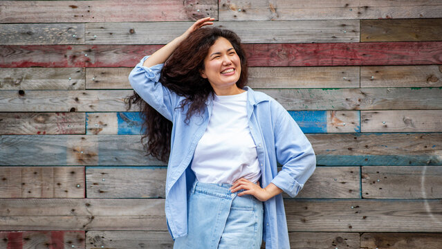 Cheerful young Asian woman smiling with hand in hair in front of rustic wooden wall