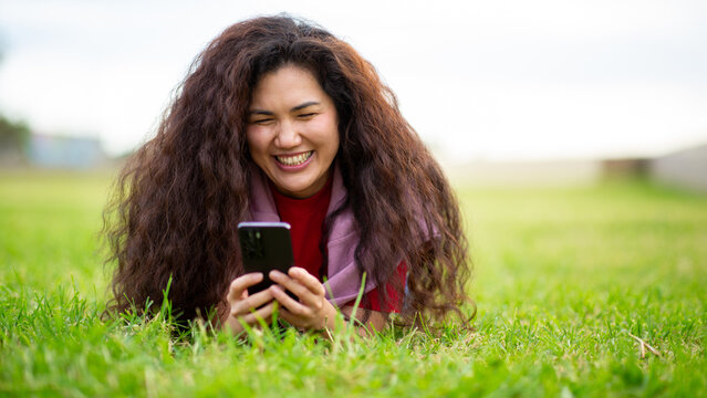 Laughing Asian woman using smartphone while lying on grass in outdoor park - Powered by Adobe