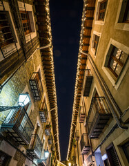 Obraz premium calle estrecha de un casco antiguo, iluminada con luces navideñas cálidas que bordean los tejados y balcones de edificios de piedra y madera, creando un ambiente festivo y acogedor durante la noche.