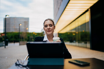 Young female professional smiling while using tablet outdoors, sipping coffee, working on business tasks in digital environment, multitasking confidently at modern urban location