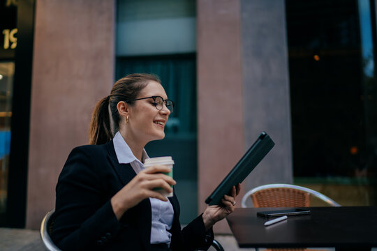 Smiling woman in business suit and glasses working on tablet while drinking coffee, sitting outside, enjoying balance between digital productivity and personal comfort