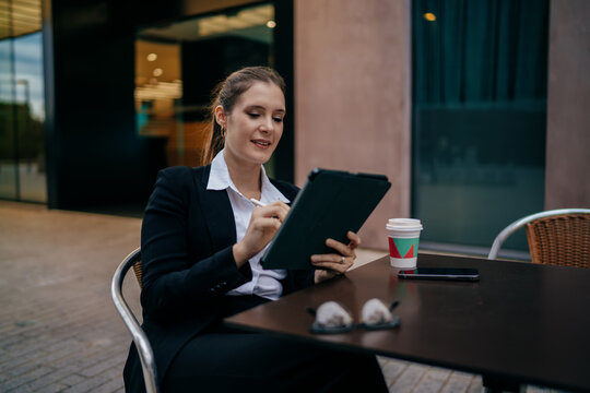 Smiling young woman in business suit using digital pen and tablet at cafe table outdoors, takeaway coffee and smartphone beside her, enjoying flexible remote work with confidence