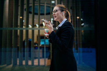 Caucasian woman in glasses and business suit sending voice message while holding takeaway coffee, illustrating multitasking and hands-free mobile interaction in remote business routine