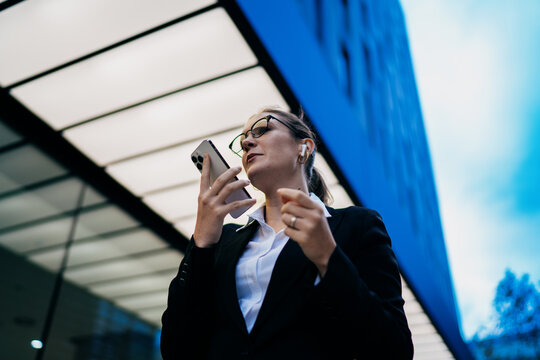 Caucasian businesswoman speaking voice command into smartphone, standing near glass facade, confidently managing business with mobile-first strategy and digital independence