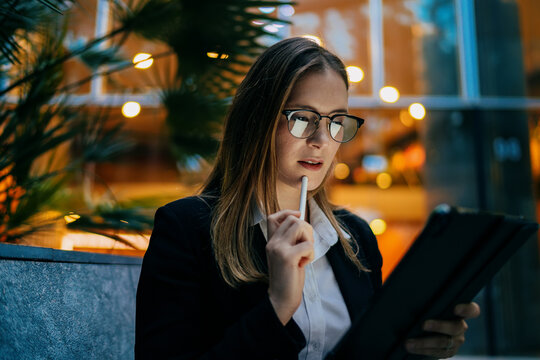 Curious woman in glasses looking at tablet while holding stylus to lips, sitting outdoors and engaging in digital learning or freelance planning after work hours