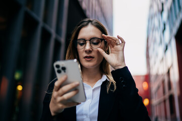 Caucasian businesswoman adjusting glasses while reading smartphone screen outdoors, focused on digital clarity and mobile workflow in smart urban routine