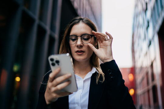 Caucasian businesswoman adjusting glasses while reading smartphone screen outdoors, focused on digital clarity and mobile workflow in smart urban routine