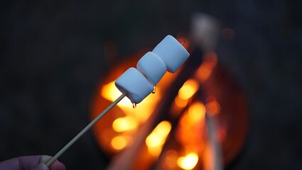 Hand holding a skewer with three marshmallows over a glowing campfire at dusk, symbolizing camping, summer fun, cozy evenings, and outdoor recreation.