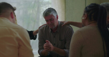 Man bows head and clasps hands during group therapy session while surrounded by peers offering...