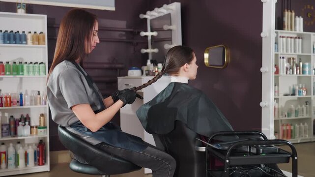 stylist braids young client's hair creating neat braid at station, gentle hands and focused technique, cape and trolley nearby, shelves of color and shampoo bottles frame modern salon atmosphere