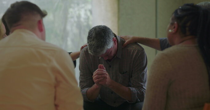 Man bows head and clasps hands during group therapy session while surrounded by peers offering support with gentle hands in quiet, caring environment