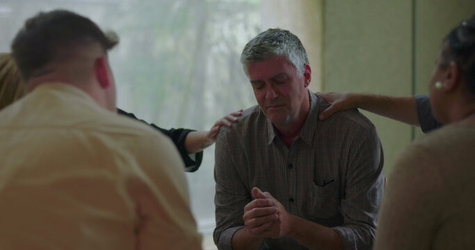 Man bows head and clasps hands during group therapy session while surrounded by peers offering support with gentle hands in quiet, caring environment