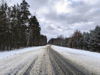 Snowy Forest Road Under Dramatic Winter Sky. Narrow snow-covered rural road disappearing into dense pine and birch forest under heavy grey clouds. Moody winter journey concept.