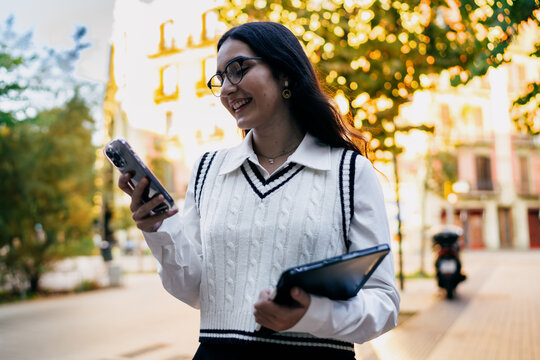Young woman uses smartphone with tablet tucked under arm, paused mid‑motion to engage digital connection, emphasising mobile tech integration in daily urban life.