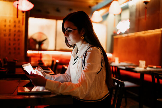 Focused young woman sitting indoors using a tablet with stylus and digital screen in a low-lit space, symbolizing multitasking, remote access, and digital communication through smart devices.