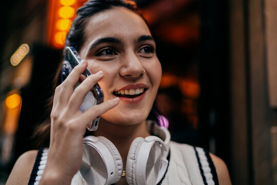 Woman with headphones around neck talks on smartphone while smiling, immersed in a friendly phone conversation, surrounded by warm evening tones and soft tech ambiance.
