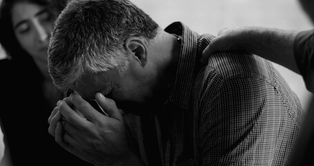 Man sobs during therapy session, burying face in hands while others gently place hands on his back...