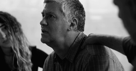 Man sobs during therapy session, burying face in hands while others gently place hands on his back and shoulder in gesture of deep emotional support and empathy