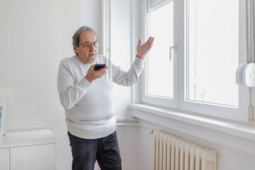 Man calls the repair service due to a problem with the cold radiator. An unhappy elderly man standing next to a cold radiator, using a cell phone to call a technical service