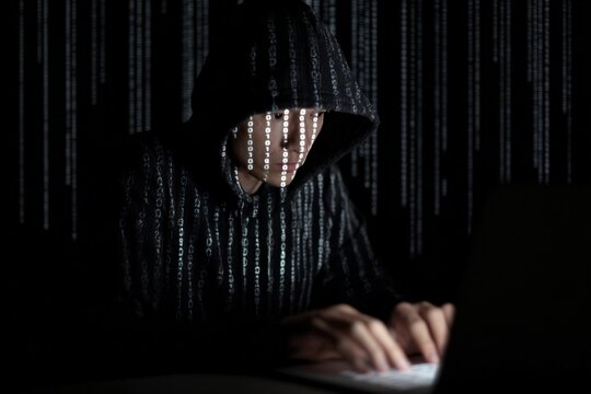 Hacker in hooded jacket typing on laptop in dark room with binary code background