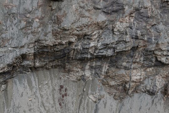 Close-up of a rough, stratified rock face jagged grey and brown layers over a wet, sandy base with small reddish specks. Natural geological textures